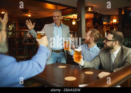 Eine Gruppe von Geschäftsleuten grüßt ihren Kollegen, während er im Café sitzt und nach der Arbeit Bier trinkt Stockfoto