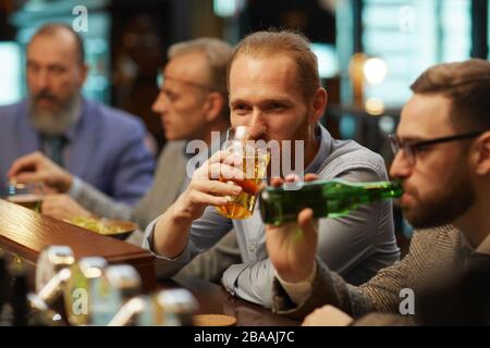 Der junge Mann genießt das Glas Bier, während er zusammen mit seinen Freunden an der Theke sitzt Stockfoto