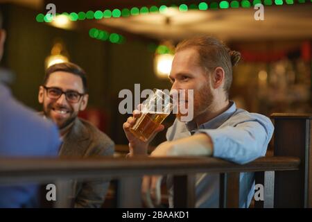 Bärtiger junger Mann trinkt Bier, während er zusammen mit seinen Freunden in der Kneipe sitzt Stockfoto