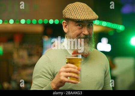 Portrait des reifen bärtigen Mannes in der Kappe, die Glas Bier hält und die Kamera in der Bar betrachtet Stockfoto