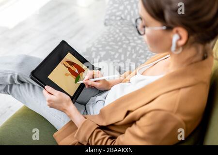 Junge kreative Frau, die als Webdesignerin oder Künstlerin arbeitet und dabei auf einem digitalen Tablet sitzt, während sie zu Hause auf dem bequemen Sofa sitzt Stockfoto