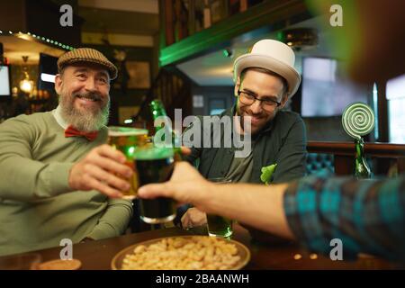 Fröhlicher reifer Mann in der Mütze, der zusammen mit seinen Freunden am Tisch in der Bar Bier trinkt Stockfoto