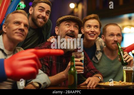 Reifer bärtiger Mann, der mit seinen Freunden ein Glas Bier am Tisch hält und in der Sportbar Fußballspiele beobachtet Stockfoto