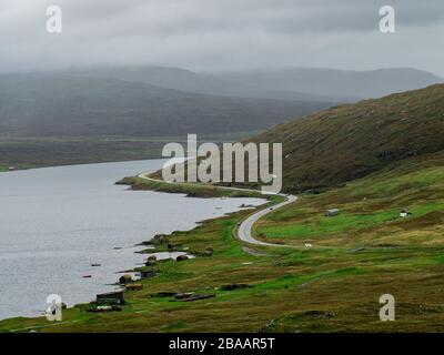 Färöer. Straße am See entlang. Grüne Felder im Vordergrund und Wolken im Hintergrund. Duftige Landschaft. Stockfoto