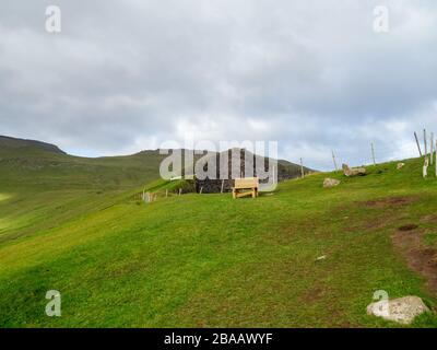 Färöer. Hänge von Hügeln, die sich über dem Dorf Gjógv erheben. Holzbank im Hintergrund. Bewölkter Himmel. Stockfoto