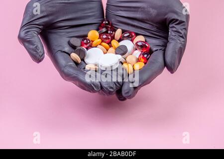 Die Hände der Männer in schwarzen Gummihandschuhen halten viele mehrfarbige Pillen auf einem rosafarbenen Hintergrund Stockfoto