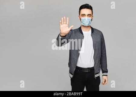 Junger gutaussehender Mann in schützender Gesichtsmaske, der mit der Hand Halt macht, isoliert auf dem grauen Hintergrund, Kopierraum. Konzept von Gesundheit und Grippe Stockfoto