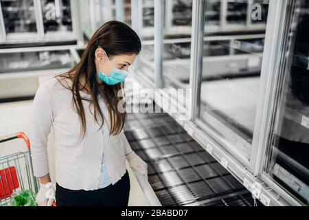 Frau mit Maske und Handschuhen kauft Lebensmittel/Verbrauchsmaterial im Supermarkt mit ausverkauften Produkten.Lebensmittelmangel.Leere Regale wegen neuartiger Korona Stockfoto