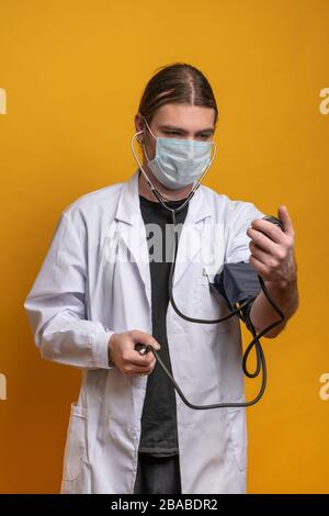 Der junge Arzt misst seine Spannung mit einem Messgerät, während er eine Schutzmaske gegen den sars-cov-2-Virus trägt. Aufnahme im Hochformat gegen oder Stockfoto
