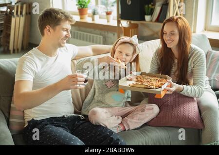 Die Familie, die zu Hause eine schöne Zeit verbringt, sieht glücklich und fröhlich aus. Mutter, Vater und Tochter haben Spaß, essen Pizza, entspannen beim Sitzen auf dem Sofa. Zusammenleben, Wohnkomfort, Liebe, Beziehungskonzept. Stockfoto