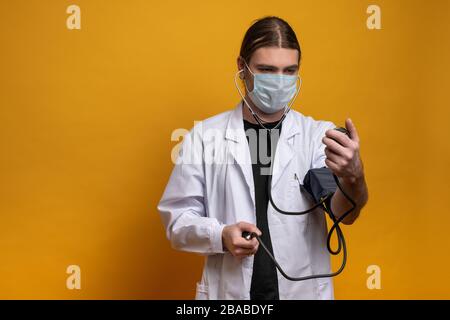 Der junge Arzt misst seine Spannung mit einem Messgerät, während er eine Schutzmaske gegen den sars-cov-2-Virus trägt. Aufnahme im Querformat gegen o Stockfoto