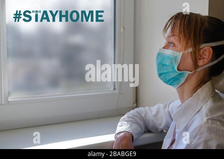Eine Frau trägt eine medizinische Maske, die vor einem Fenster mit der Aufschrift "nach Hause" auf einem verschwommenen Stadthintergrund sitzt. Bleiben Sie zu Hause, rufen Sie während des COVID-1 an Stockfoto