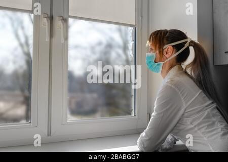 Traurige Frau in einer Maske, die ins Fenster schaut. Selbstisolation zur Verhinderung einer Pandemie des Corona-Virus-Kovid-19. Stockfoto