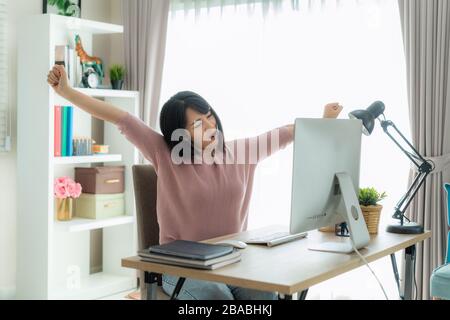 Asiatische Geschäftsfrau arbeitet von zu Hause aus und streckt ihren Körper aus, weil sie müde und schläfrig ist, nachdem sie an einem Computer gearbeitet hat, eine intelligente Frau, die zu Hause arbeitet. Leben Stockfoto