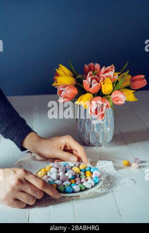 Männliche Hände legen Teller mit Ostern bunten süßen Eiern auf den weißen Holztisch mit frischer Tulpenblumen Vase und dunkelblauem Hintergrund. Familie Holi Stockfoto