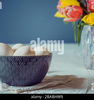 Frische weiße Eier in keramisch-grauer Schüssel auf Vintage-Serviette und Vase mit frischen rosa und gelben Tulpen auf dem weißen Holztisch und dunkelblauem Hintergrund. Stockfoto