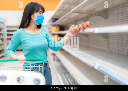 Asian Woman holt letzte Ei-Packung im Supermarkt leere Regale inmitten der COVID-19-Coronavirus Ängste, Käufer panischen Kauf und Lagerung von toilettenpapier vor Stockfoto