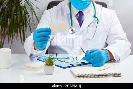 Doktor in einem weißen medizinischen Mantel sitzt an einem Tisch in einem braunen Lederstuhl und hält eine rechteckige weiße leere Visitenkarte in den Händen, medizinisches Büro Stockfoto