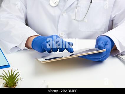 Doktor in einem weißen medizinischen Mantel sitzt an einem Tisch und hält eine elektronische Tablette in der Hand, berührt den Bildschirm mit der rechten Hand, Arztpraxis, di Stockfoto