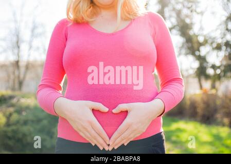 Schwangere Dame in rosafarbenem Hemd, die an einem sonnigen Frühlingnachmittag draußen steht und ihre Hände am Bauch hält Stockfoto