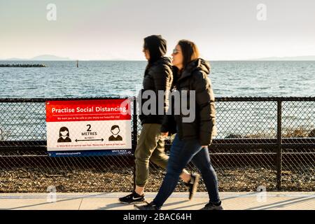 White Rock, Kanada - 25. März 2020: Zwei Fußgänger, die an der sozialen Distanzierungsbenachrichtigung Covid-19 vorbeigehen Stockfoto