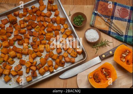 Würfel aus geröstetem Butternusskürbis in Pfanne und rohen Hälften auf Schneidebrett mit Kräutern und Gewürzen in kleinen Holzschüsseln. Stockfoto