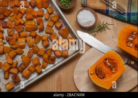 Würfel aus geröstetem Butternusskürbis in Pfanne und rohen Hälften auf Schneidebrett mit Kräutern und Gewürzen in kleinen Holzschüsseln. Stockfoto