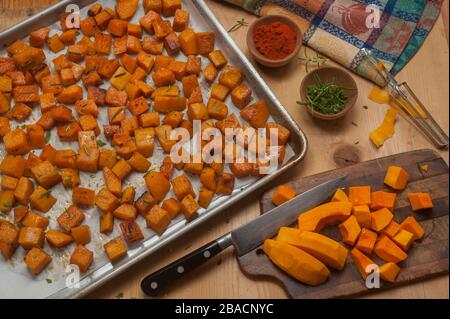 Würfel aus geröstetem Butternusskürbis in Pfanne und rohen Hälften auf Schneidebrett mit Kräutern und Gewürzen in kleinen Holzschüsseln. Stockfoto