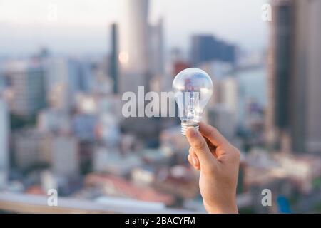 Glühbirne vor Sonnenuntergang städtischen Häuser. Glühlampe transparent Lampe in alternative Solar Energie des Menschen in der Stadt, Stadt, Stadt Nachtleben. Symbol, Konzept der Busi Stockfoto