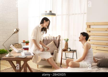 Junge Frau sitzt auf dem Schreibtisch im Schlafzimmer und ihr Mann chattet Stockfoto