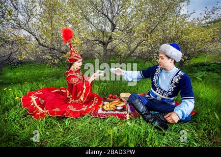 Paar im kasachischen Kostüm sitzt im Frühjahr blühen Apfelgarten bei Picknick und Teetrinken in Almaty, Kasachstan, Zentralasien Stockfoto