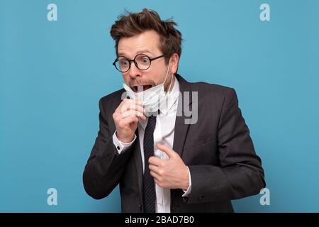 Der junge gutaussehige Mann in medizinischer Maske hat Panikattacke. Er fürchtet, dass er infiziert ist. Studio an blauer Wand gedreht Stockfoto