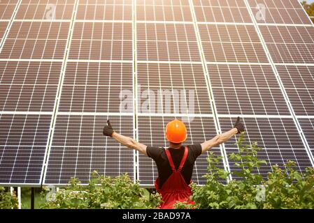 Arbeiter, der mit dem Rücken zur Kamera vor dem Hintergrund großer Plantagensolarbatterien steht und Daumen zeigt, wie Zeichen. Hausbau. Grüne ökologische Energieerzeugung. Stockfoto
