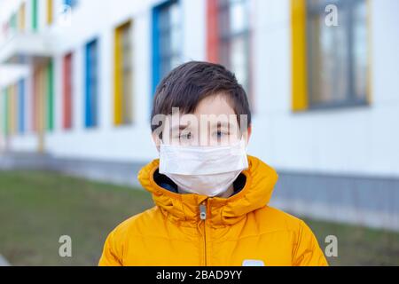 Portrait eines Jungen in einer gelben Jacke in einer weißen medizinischen Maske auf der Straße in der Nähe des Gebäudes mit mehrfarbigen Fenstern.Concept Disease Quarantäne Stockfoto