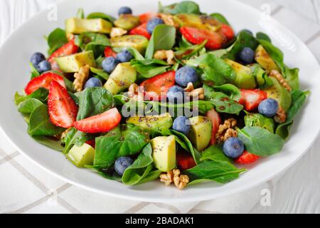 Nahaufnahme von frischem Spinat Avocado-Walnüssen Erdbeere und Blaubeersalat mit Balsamico-Essig-Olivenöl und Mohnsamen, die auf einem weißen Teller anstehen Stockfoto