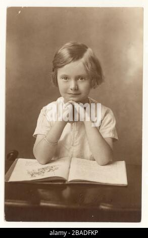 Anfang 1900 erschien das Studio-Porträt eines süßen jungen Mädchens mit typischer Frisur, das mit einem offenen Buch sitzt, das um die 1930er Jahre in Großbritannien entstand Stockfoto