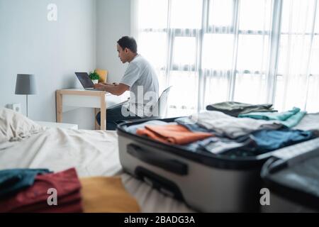 asiatischer junger Mann überprüft die Abfahrtszeiten per Online-Bewerbung, nachdem er sie vorbereitet hat und legt ihre Kleidung in den Koffer Stockfoto