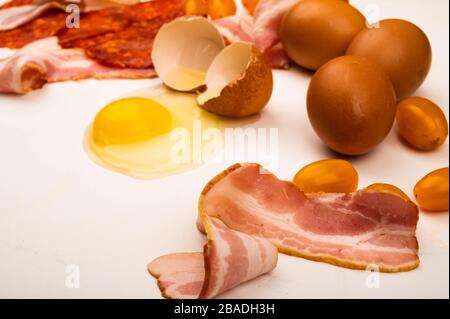 Zerbrochenes Hühnerei, Speck- und Wurstscheiben sowie Tomaten auf weißem Hintergrund. Nahaufnahme Stockfoto