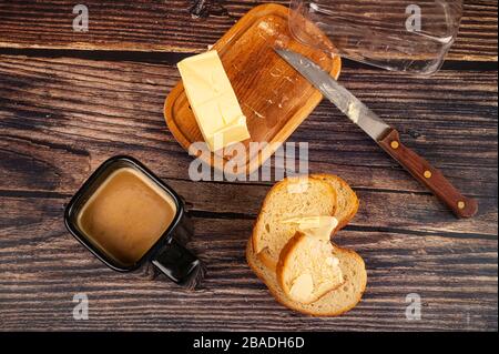 Frischer Weizentoast mit Butter, Butter in einer Holzbutterform, ein Messer mit Holzgriff und eine Tasse Kaffee mit Milch auf Holzgrund. Clos Stockfoto