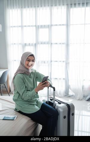 Junge asianerin sitzt auf dem Bett und hält ihr Handtelefon und ihren Koffer, bevor sie in den Urlaub geht Stockfoto