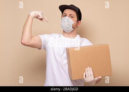 Junger Auslieferer in medizinischen Masken, der eine cardbox hält und trägt, die Bizeps zeigt, die auf beigem Hintergrund isoliert sind Stockfoto