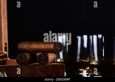 Stillleben mit drei kubanischen Zigarren, zwei Gläsern Whisky oder Rum und Holzkiste mit Hygrometer und schwarzem Hintergrund. Platz für Ihren Text. Stockfoto