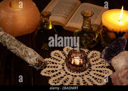 Esoterisches Stillleben mit brennender Kerze in gelber Flamme, offenem alten Buch und Kristallen. Zwei grüne Fläschchen und getrockneter Salbei auf Holztischplatte. Stockfoto