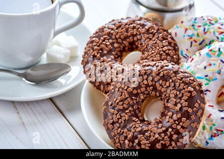 Nach oben Closeup Foto mit süßem Frühstück oder Snacks. Schokolade und Vanilledonuts und Tasse schwarzen Kaffee auf weißem Holztisch. Stockfoto