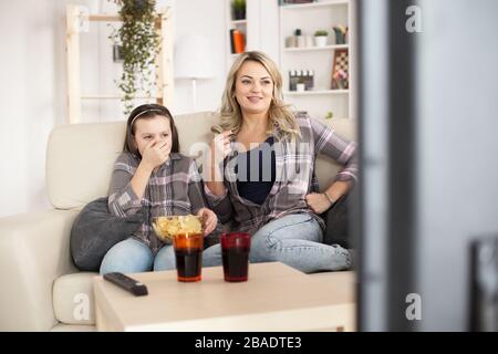 Junge Mutter und ihr kleines Mädchen sehen sich Filme im großen Fernsehen im Wohnzimmer an, essen Chips und trinken Limonade. Stockfoto