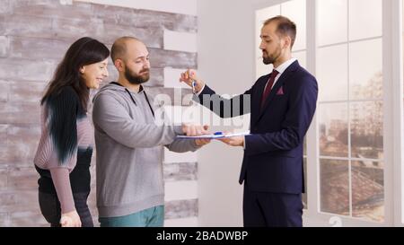 Junge Paare werden Hausbesitzer, nachdem sie Dokumente mit Immobilienmakler in Geschäftsanzug unterschrieben haben. Agent gibt Schlüssel zum Paar. Stockfoto
