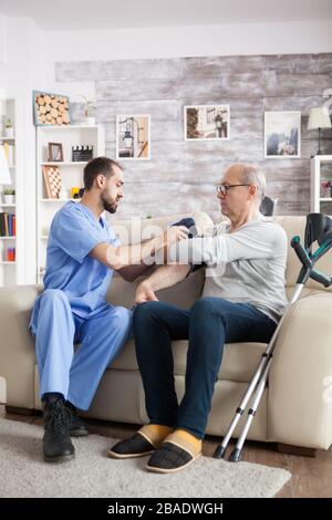 Junger Assistent im Pflegeheim überprüft den Blutdruck des alten Mannes, der auf der Couch sitzt. Stockfoto