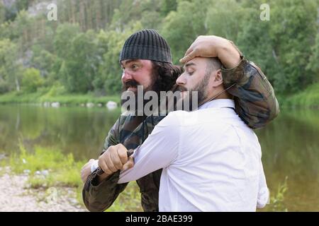 Brutaler bärtiger Terrorist in Tarnung hält langes Messer am Hals des jungen verängstigten Mannes in weißem Hemd am Ufer des Sees. Geiselnahme, Terroristendreier Stockfoto