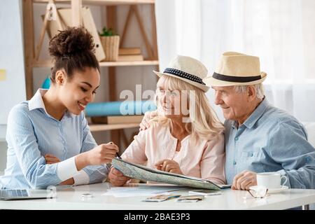 Reisebüro im Büro zeigt den älteren Kunden das Reiseziel in Hüten, die über die Wahl diskutieren Stockfoto