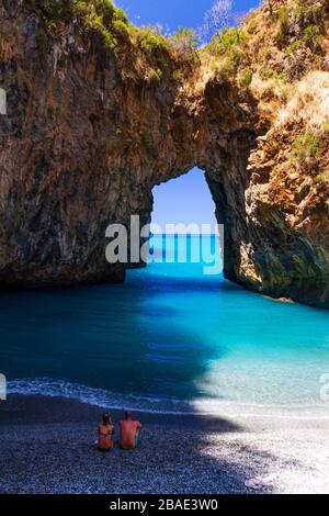 Frau und Mann sitzen an einem Kieselufer und beobachten das Meer, an einem Ort neben Tropea, genannt "Arco Magno" Stockfoto